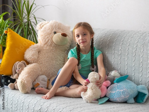 A little girl is sitting on the couch with a big teddy bear and other soft toys, smiling. The room is a mess. A lot of things are scattered. Modern interior with yellow curtains