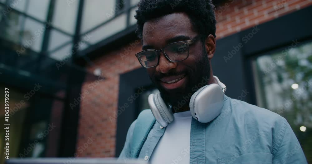 Camera moving closer to African American man standing outside under modern building. Using large tablet device. Typing on screen with one hand. Wearing headphones on his neck. Smiling. Camera moving closer to African American man standing outside under modern building. Using large tablet device. Typing on screen with one hand. Wearing headphones on his neck. Smiling.