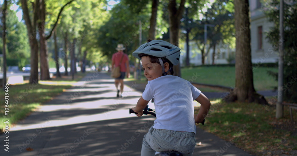 Fototapeta premium Young boy riding a bicycle with a helmet, glancing back with a playful expression, biking along a shaded park path during summer, enjoying childhood freedom and outdoor exploration