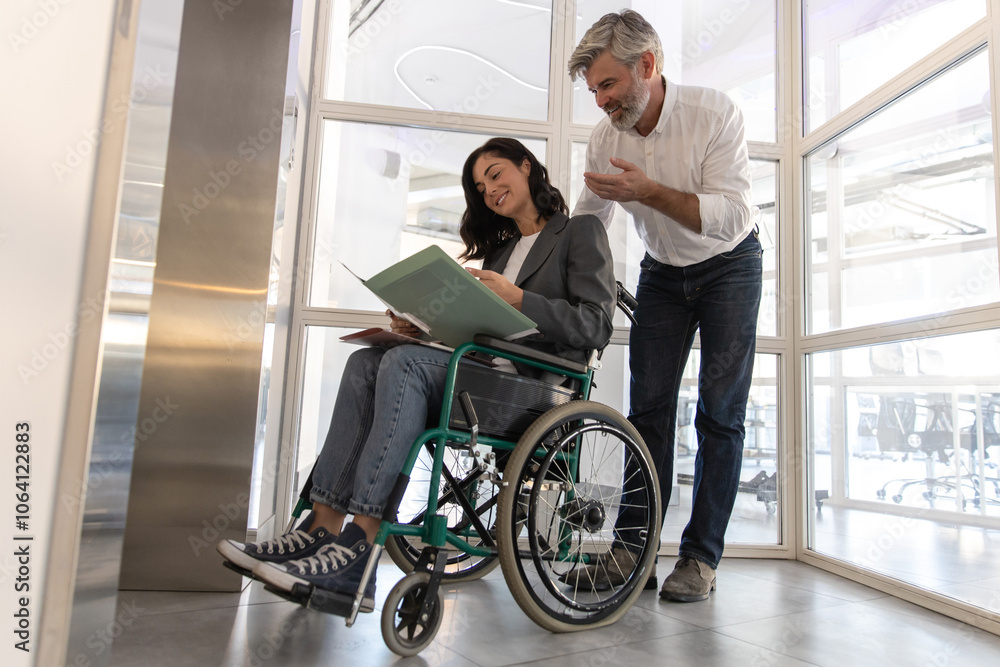 Man communicating with female in wheelchair near list in office hall Man communicating with female in wheelchair near list in office hall