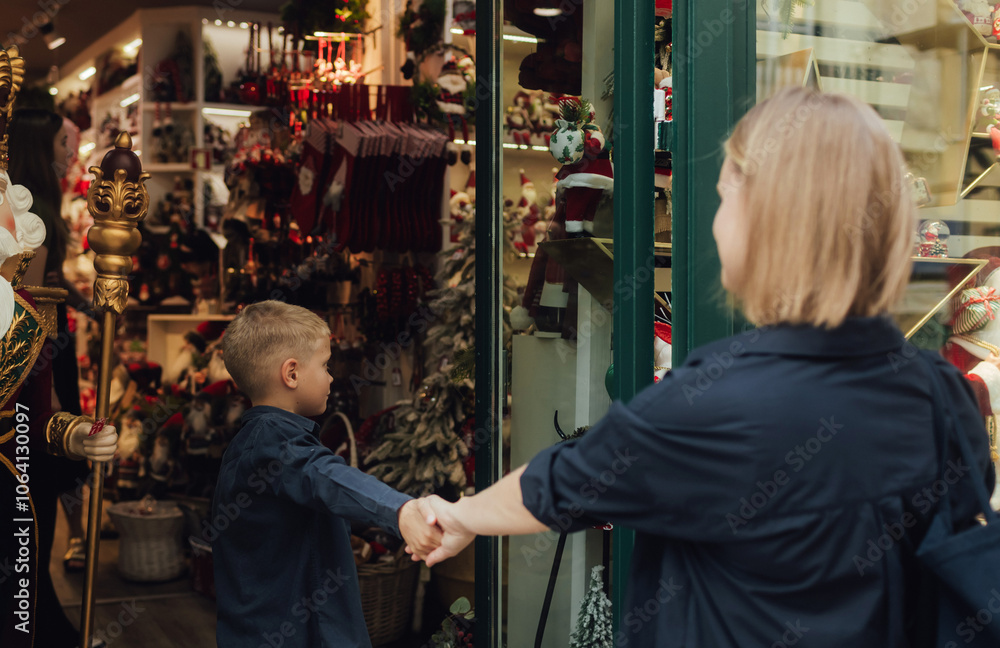 Cute little school kid boy with mother on Christmas market. Mom and son look in the window of a gift shop and choose surprises for the family. Holidays, Christmas, childhood and people concept Cute little school kid boy with mother on Christmas market. Mom and son look in the window of a gift shop and choose surprises for the family. Holidays, Christmas, childhood and people concept