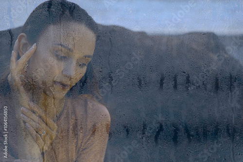 close-up of raindrops on glass, with a blurred womans face in front. dreamy female with sad eyes closed, water rain droplets flow near head, condensat against a refreshing background. rainy weather.