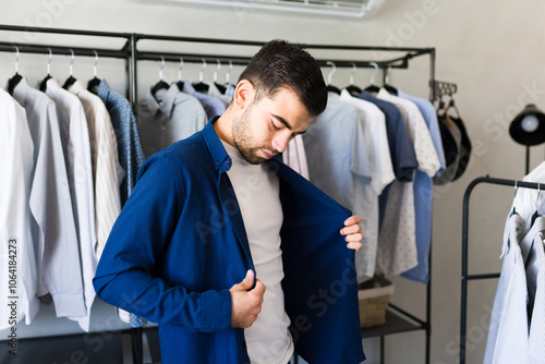 Man standing in a dressing room surrounded by clothes, trying on a stylish blue shirt