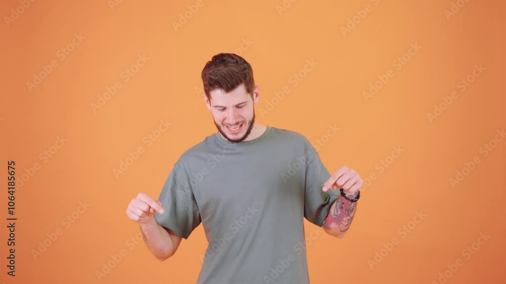 A young man with a tattoo on his arm points downward with confidence, directing attention below him. He stands against a vibrant orange background, looking focused A young man with a tattoo on his arm points downward with confidence, directing attention below him. He stands against a vibrant orange background, looking focused