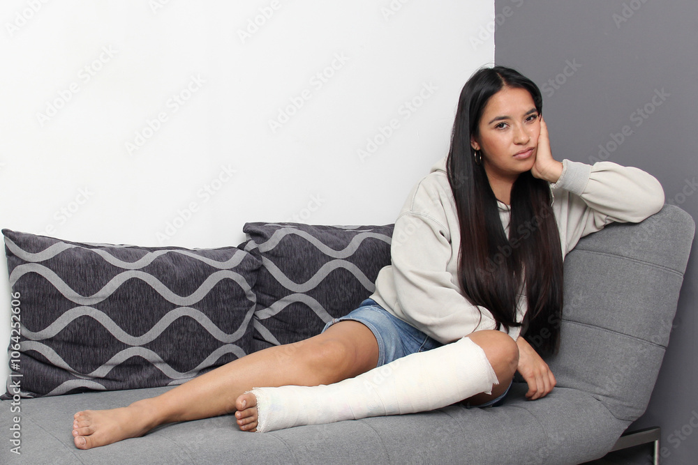 Young 30-year-old woman is sad in her living room because she broke her leg and has it in a cast without being able to move Young 30-year-old woman is sad in her living room because she broke her leg and has it in a cast without being able to move