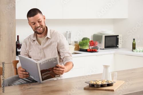 Young man with tasty muffins reading magazine in kitchen