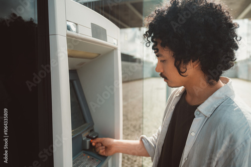 A wide-angle shot of a young man with curly hair and a mustache stands at an outdoor ATM, focused on the screen as he inserts his bank card. Dressed casually in a light shirt and dark t-shirt photo