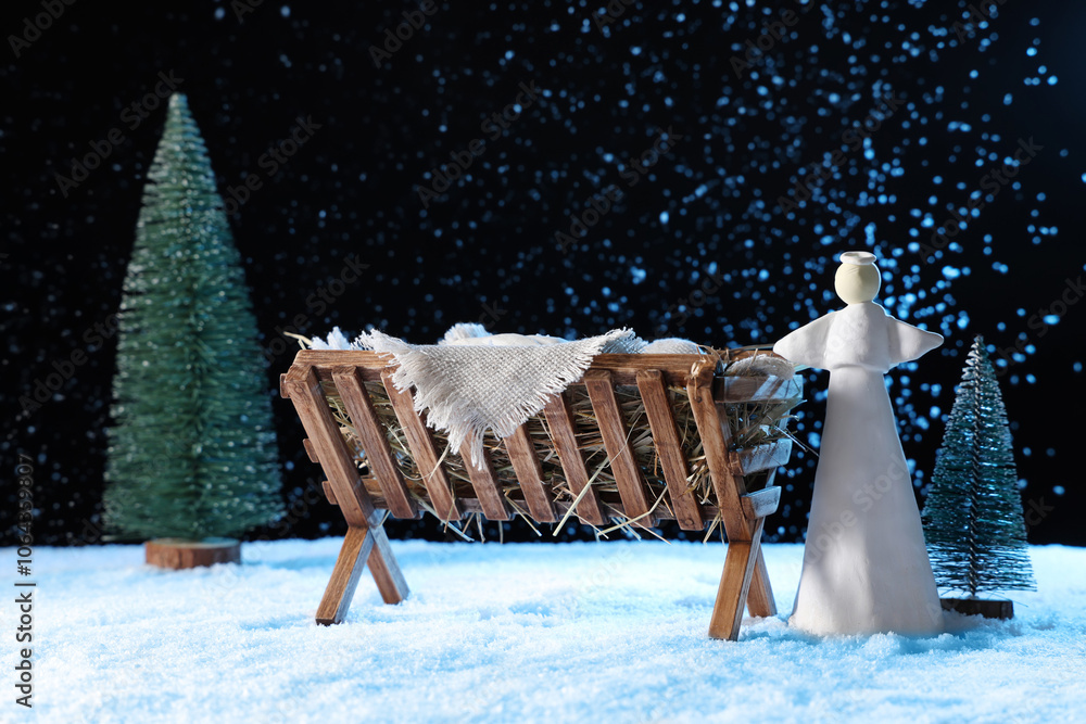 Naklejka premium Manger with baby Jesus, angel and Christmas trees on snow against dark background