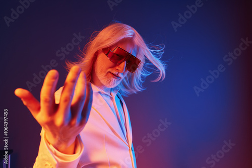 Stylish older man with long hair wearing oversized sunglasses and a white suit, posing confidently against a colorful gradient background