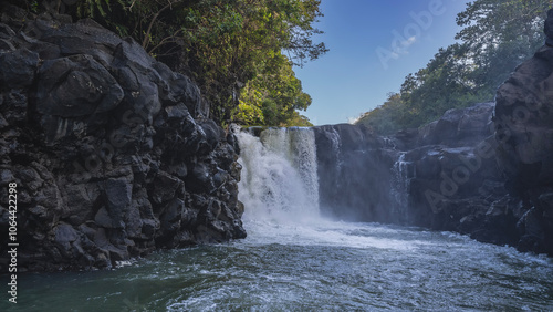 Fototapeta Naklejka Na Ścianę i Meble -  A beautiful tropical waterfall. Streams flow down from the cliff ledge into the river. Splashes, foam. Green vegetation on the rocks. The blue sky. Mauritius. Grand River South East waterfall 