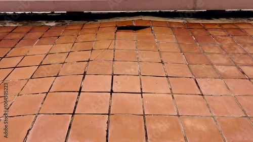 A distressed and uneven outdoor tile floor, showing significant damage and dislodged tiles. The image captures the effects of weathering and wear, emphasizing structural deterioration and maintenance 