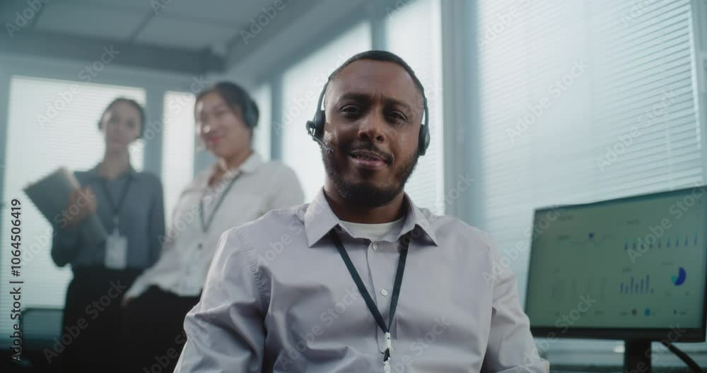 Call center office: Close up portrait of friendly African American technical customer support operator wearing headset smiling, looking at camera. Team of helpdesk specialists standing in background. Call center office: Close up portrait of friendly African American technical customer support operator wearing headset smiling, looking at camera. Team of helpdesk specialists standing in background.