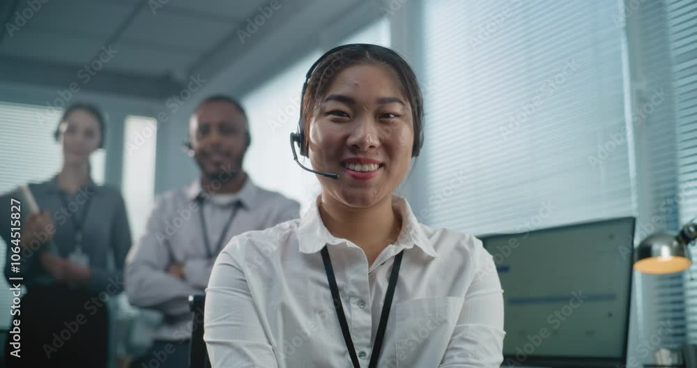 Call center office: Close up portrait of smiling Asian female technical customer support specialist in headset looking at camera. Unfocused multiethnic team of hotline operators in the background. Call center office: Close up portrait of smiling Asian female technical customer support specialist in headset looking at camera. Unfocused multiethnic team of hotline operators in the background.