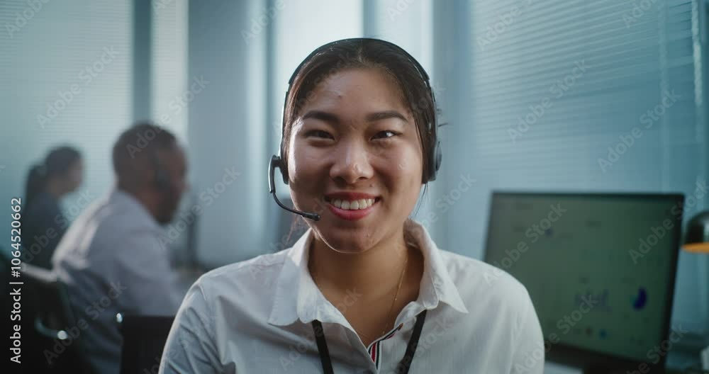 Call center office: Close up portrait of friendly Asian female technical customer support specialist wearing headset smiling and looking at camera. Team of hotline operators working in the background. Call center office: Close up portrait of friendly Asian female technical customer support specialist wearing headset smiling and looking at camera. Team of hotline operators working in the background.