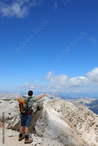 young hiker with a backpack on his shoulders admiring the view pointing at the peaks of the dolomites in the european alps