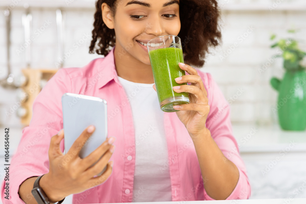 Morning Smoothie. Closeup portrait of african woman using phone and drinking homemade detox juice in kitchen Morning Smoothie. Closeup portrait of african woman using phone and drinking homemade detox juice in kitchen