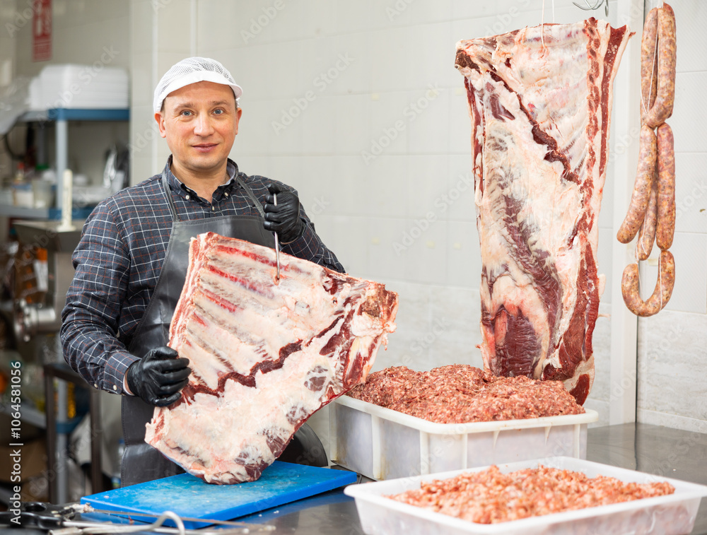 Φωτογραφία Adult male butcher holding beef ribs hanging on hook