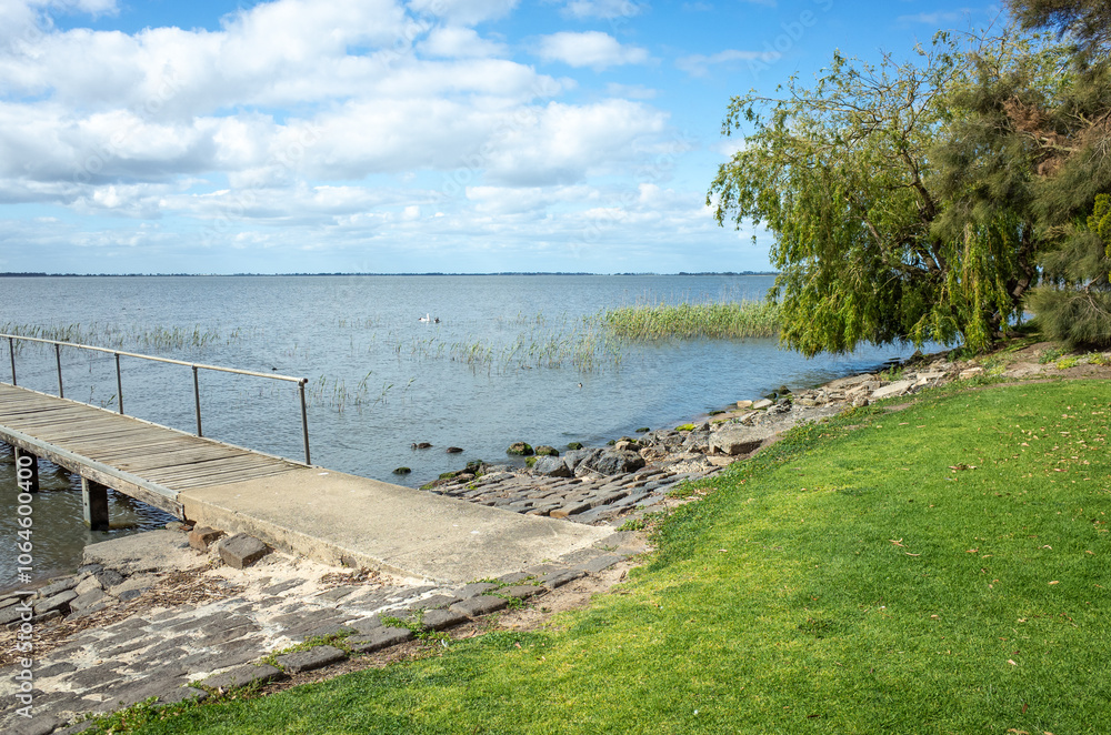 Fototapeta premium A picturesque lakeside scene at Lake Colac, Australia with a wooden dock or pier extending out over calm water. The shoreline is lined with lush, green grass and a weeping willow tree.