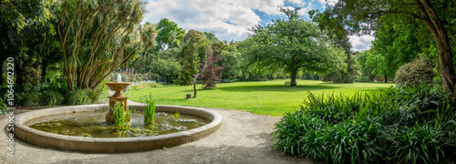 Fotografie Panoramic garden view with a circular fountain in an ornamental pond, lush landscaping, and a manicured lawn