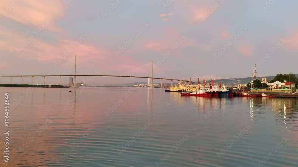 Serene dawn over Cebu Cordova Link Expressway Bridge, Philippines, with soft sky hues reflected on calm peaceful water as industrial boats await at docks Serene dawn over Cebu Cordova Link Expressway Bridge, Philippines, with soft sky hues reflected on calm peaceful water as industrial boats await at docks
