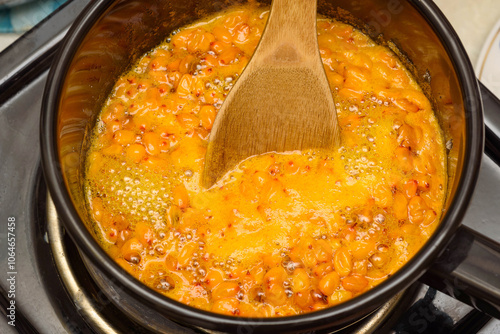 Stirring boiling sea buckthorn berries with sugar in a saucepan with a wooden spoon.