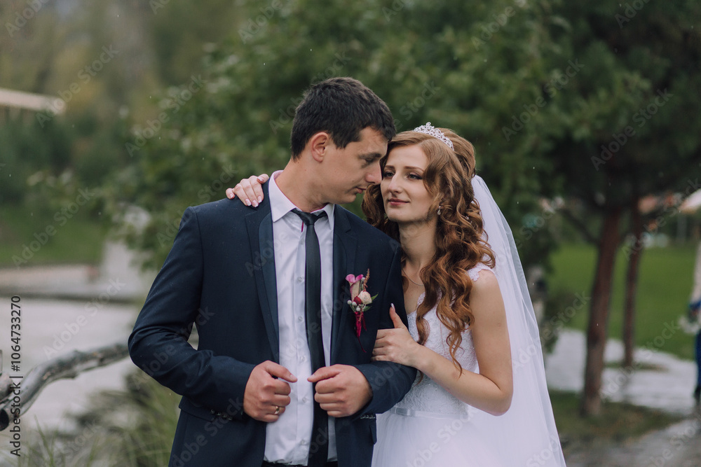 A bride and groom are standing in the rain, with the bride wearing a veil and the groom wearing a tie A bride and groom are standing in the rain, with the bride wearing a veil and the groom wearing a tie