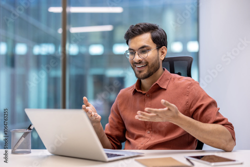 Smiling Hispanic businessman sitting at desk using laptop for video call in modern office setting. Glasses and professional attire emphasize business context. Concept of communication, technology