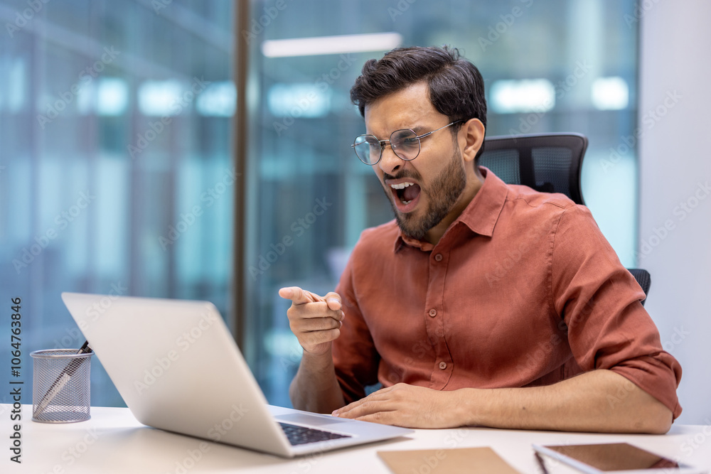 Business man showing frustration while pointing at laptop in office. Expresses emotions like anger or dismay during work. Seated at desk with stationery in modern environment. Business man showing frustration while pointing at laptop in office. Expresses emotions like anger or dismay during work. Seated at desk with stationery in modern environment.