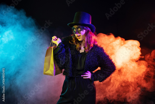 Young woman wearing black hat, mask, clothes holds paper bag on darkness background with magical colorful smoke. Black friday shopping or sales.