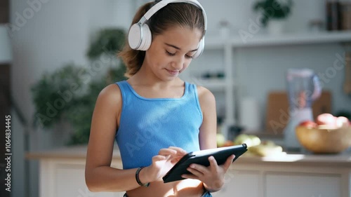 Video of beautiful little girl with headphones while using digital tablet standing in the kitchen