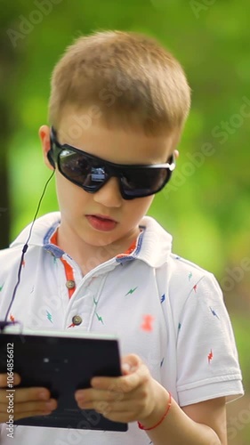 A boy walks in the park and listens to music on headphones on a tablet. Vertical video.