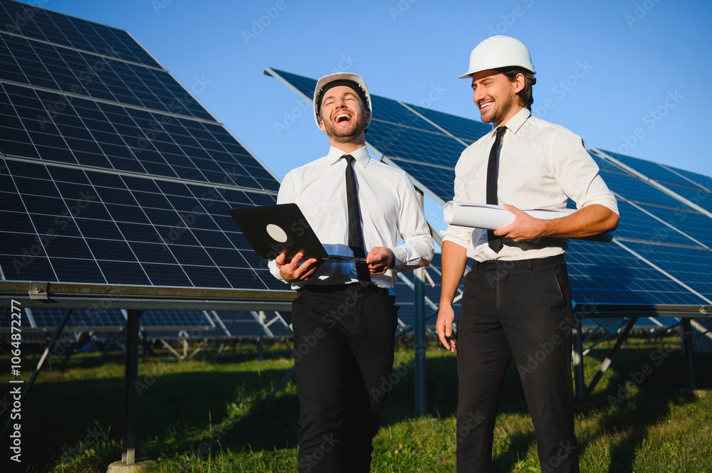 Focused male engineers in hardhats using laptop and discussing project while standing near photovoltaic batteries in countryside Focused male engineers in hardhats using laptop and discussing project while standing near photovoltaic batteries in countryside