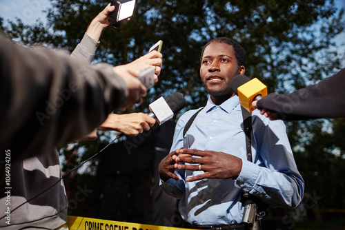 Low angle portrait of young African American man as police officer talking to reporters on crime scene and speaking to multiple microphones with camera flash photo