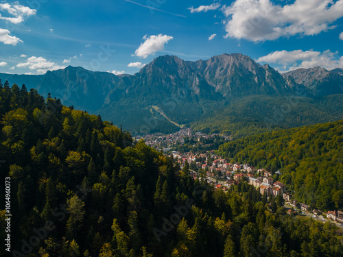 A town at the base of the mountain in the autumn season. Astonishing aerial view with the rocky mountains filmed in cinematic style. Romanian landscape above Busteni city in a sunny day with blue sky