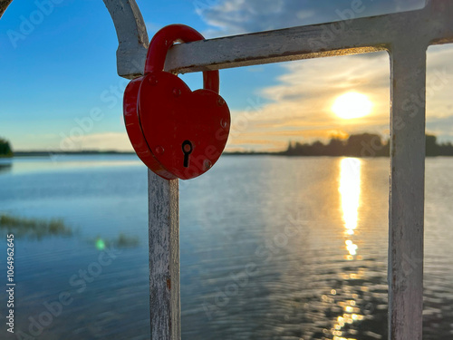 A red heart padlock is attached to a railing by a lake