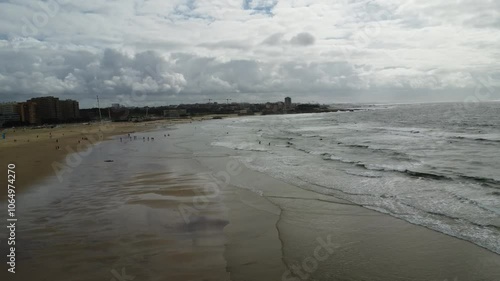 vista aérea da praia de Matosinhos capturada por um drone. O mar azul profundo se estende até o horizonte, com ondas suaves que acariciam a extensa faixa de areia dourada. Na praia, pequenos pontos co photo