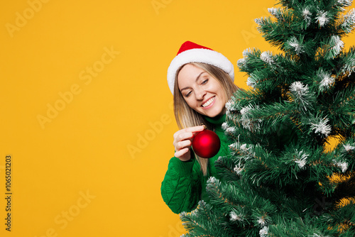 Full body merry smiling young woman wear green casual sweater Santa hat posing stand behind Christmas tree decorate with bauble toy isolated on plain yellow background Happy New Year, holiday concept