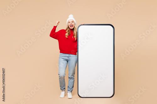 Full body merry young woman wear red sweater hat big huge blank screen area mobile cell phone smartphone do winner gesture isolated on plain beige background. Happy New Year Christmas holiday concept. photo