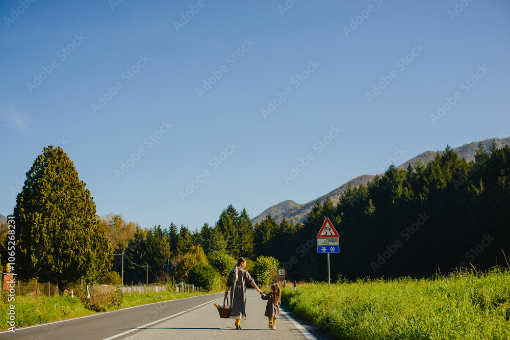 Obraz premium Mom and daughter walk along the road by the hand against the background of the forest and mountains. Mom and a girl in dresses. There is a picnic basket in his hands. Italian alps, countryside, tranqu