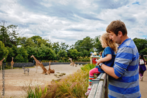 Cute adorable toddler girl and father watching and feeding giraffe in zoo. Happy baby child, daughter and dad, family having fun together with animals safari park on warm summer day. Ireland