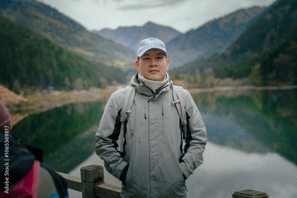 Fototapeta premium Hiker man wearing jacket and backpack standing by the mountain lake and looking at camera, Dagu Glacier, Chengdu, China.