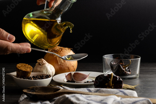 Una mano sostiene una jarra de aceite vertiendo sobre una cuchara sopera. Alrededor, dientes de ajo, ñoras, cebolla y bolas de pimienta crean una escena vibrante, resaltando la riqueza de los ingredie photo