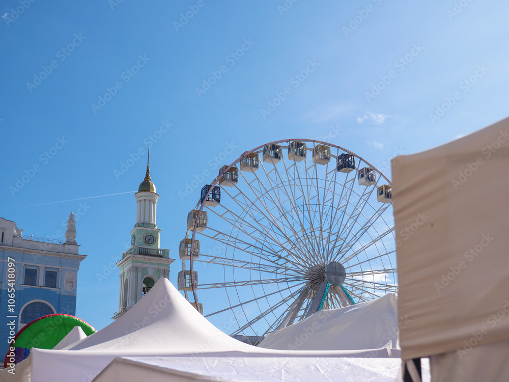 Modern observation wheel and ancient church bell tower with bright blue cloudless sky in the background in Kontraktova square, Podil, Kyiv city. Travelling and entertainment in old city center.