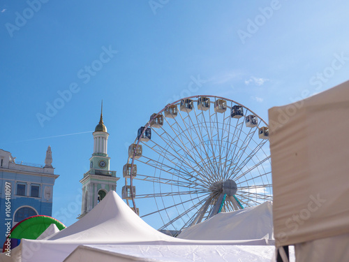 Obraz na płótnie Modern observation wheel and ancient church bell tower with bright blue cloudless sky in the background in Kontraktova square, Podil, Kyiv city. Travelling and entertainment in old city center. w salonie