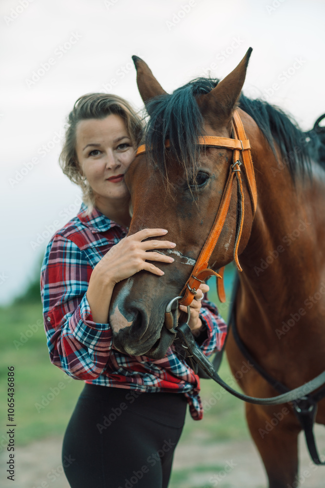 Obraz premium Happy blonde with horse in forest. Woman and a horse walking through the field during the day. Dressed in a plaid shirt and black leggings.