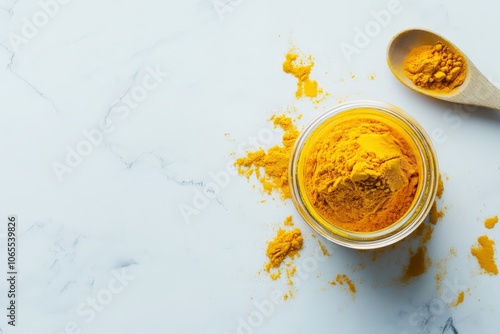 Turmeric powder, strikingly vibrant and yellow, sprawls around an open glass jar on a marble surface, emphasizing its culinary and health benefits vividly. photo