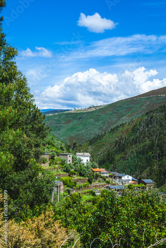 Wallpaper Mural A scenic view of a small, secluded village nestled among green hills under a bright blue sky with fluffy white clouds Torontodigital.ca