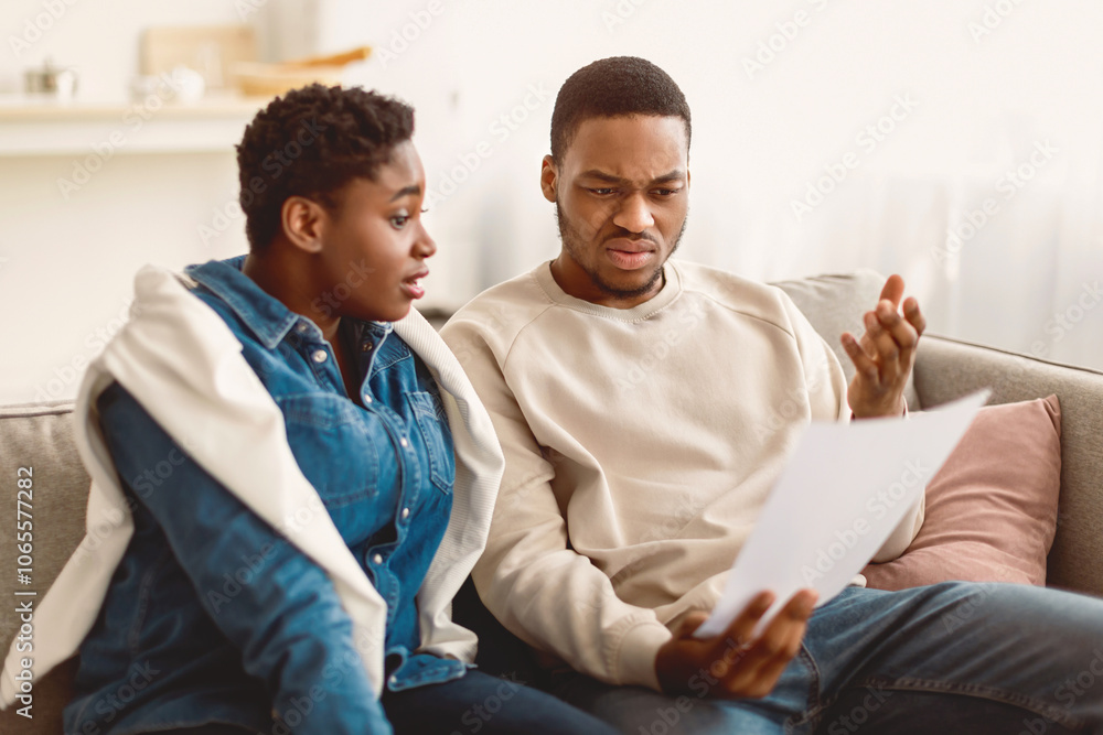 Obraz premium Financial Crisis Concept. Portrait Of Confused Perplexed African American Man Holding Paper Document And Reading Bank Notification Or Eviction Notice, Scared Woman Sitting On The Sofa In Living Room