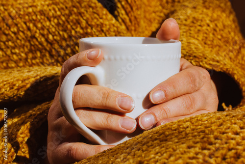 White cup in female hands. Woman wrapped in yellow plaid is drinking hot- tea or coffee. Good morning. Autumnal cozy sunny day. Relax, leisure mood.