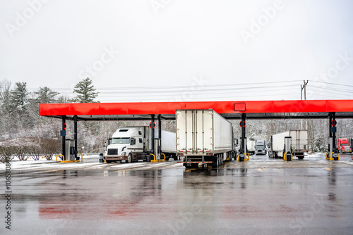 Industrial grade different Long haul big rig semi trucks with semi trailers standing on the truck stop gas station fill up the tanks to continue the route at winter time with snow and ice